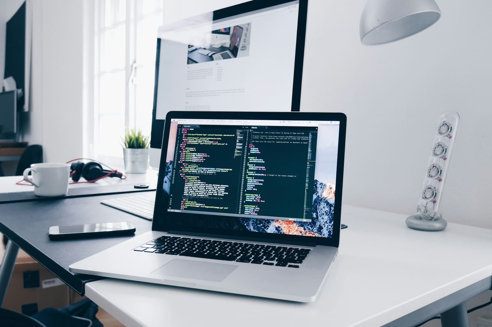 A bright desk with a laptop open on a code editor, a monitor behind it and a coffee cup to one side